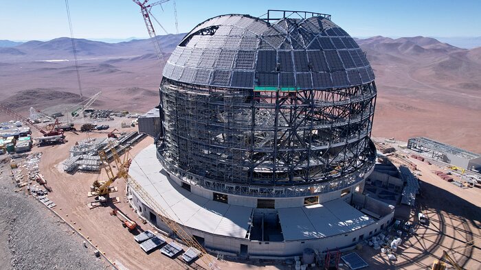 This image shows a dome structure under construction atop a mountain, against blue skies and a desert landscape. The top of the dome has been cladded, whilst towards the bottom, the metallic skeleton of the dome is still exposed. There's construction equipment and vehicles in the background of the image.