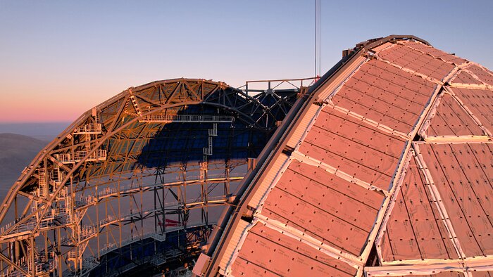 This image shows a close-up of the top of a cladded dome with a slice missing from the centre. The external cladding of the dome is tinged pink from the sunset in the background. The opening of the dome reveals its metallic skeleton inside.