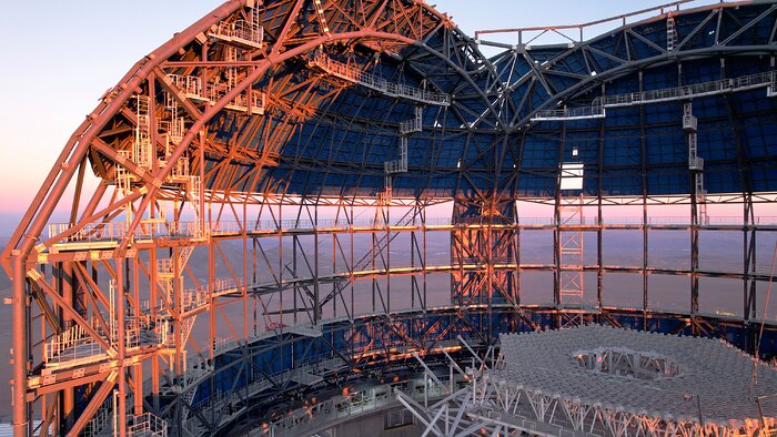 This image shows a close up of the top of a metallic dome structure, with a slice missing. The outside of the dome is fully cladded, whilst within, the skeleton of the dome is exposed. The image is tinged pink by the sunset in the background.