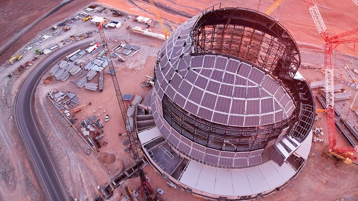 This image shows an aerial view of a metallic dome atop a desert mountain. The dome has a slice missing from its roof, and is surrounded by construction equipment, vehicles, and cranes.