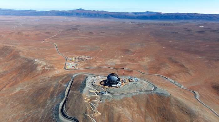 This image shows a metallic dome on top of a mountain, against blue skies and the landscape of the Chilean desert. Cranes and other construction equipment are around the dome. At the bottom of the mountain there is a group of low buildings, resembling a very small town. Towards the left of the image, there are several white buildings. In the far background, there are some very small observatories perched on a distant mountain.