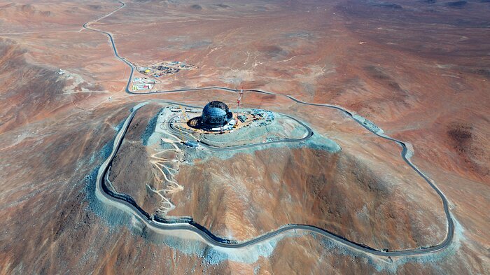 This image shows a metallic dome on top of a mountain, against the landscape of the Chilean desert. There are cranes and other construction equipment around it, as well as a road winding down the mountain leading to a collection of buildings.