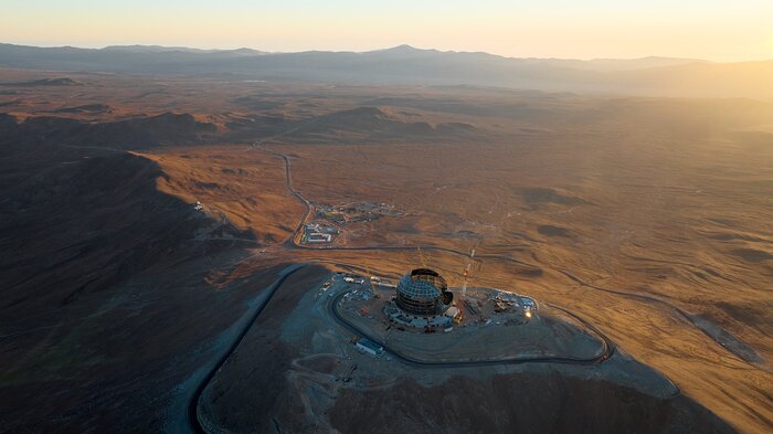 This image shows a metallic dome on top of a mountain, against the landscape of the Chilean desert at sunset. There are cranes and other construction equipment around the dome. At the bottom of the mountain there is a group of low buildings, resembling a very small town. In the far distance, a silhouette of a mountain range can be seen, and three very small observatories perched on top.