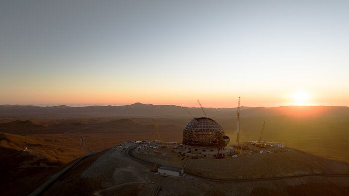 This image shows a metallic dome sat atop a mountain, against a brilliant desert landscape at sunset. In the bottom left, several distant white buildings are visible. In the mountains in the background there small silhouettes of several telescopes perched on them.