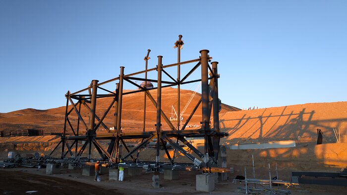 In this image, a large, metallic beam structure is shown in the foreground. Several men are working on the lower part of the structure. In the background there's a metallic dome on top of a mountain in a desert landscape.