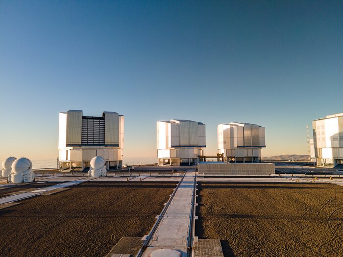 The image shows the platform on the top of the mountain which hosts the VLT. Each Auxiliary Telescope on the left has a spherical dome that lies on a cylindrical base. The Unit Telescopes are bigger and cylindrically-shaped. All the telescopes are whitish in colour. From left to right, they are arranged as follows: two Auxiliary Telescopes one behind the other, then an Auxiliary Telescope and behind the UT1, further on the right UT2 and UT3, and in the right part of the picture there is UT4, partly out of the picture. The sky is clear blue, and it gets more orange towards the horizon.