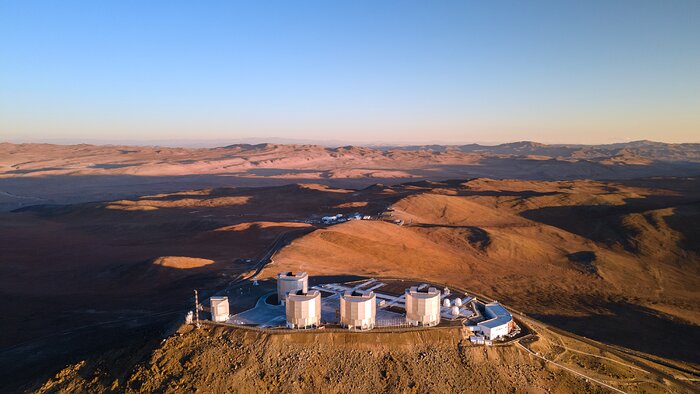 Central in the image is the VLT platform which sits on top of the Cerro Paranal mountain. On the platform, four large, cylindrical structures can be seen. These are the Unit Telescopes. On the right of the platform, three spherical white structures are just visible. These are the Auxiliary Telescopes. Beyond the platform lies the mountainous Atacama Desert, stretching off into the distance. The sky is blue at the top of the image, although it appears redder at the horizon as the Sun is setting out of shot to the right. On the left of the image long shadows are cast by the mountains, while their right hand faces appear golden in the evening Sun.