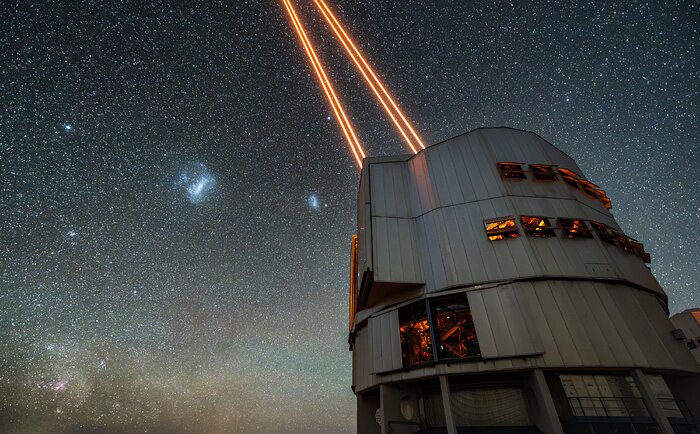 On the right of the image is the Unit Telescope 4, a cylindrical-shaped building with some windows open. From the top of the telescope, two pairs of orange lasers go up through the sky. The night sky background is full of whitish stars, and it is blue and darker in the highest part of the picture, and greenish and a bit lighter in the lowest part.