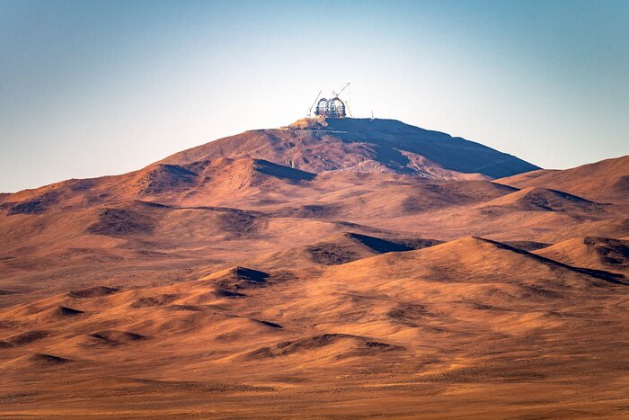 The Martian landscape of the Atacama Desert in this image is breathtaking. Under the glow of the evening sunlight, which is streaming in from the left hand side, the ground adopts a brown and red hue. The lumps and bumps of this mountainous landscape cast shadows on the ground. Mountains fill the majority of this image; climbing up the frame, we reach the flat mountain top of Cerro Armazones, positioned just a bit right of centre in the image. There we find the criss-cross steel structure of the ELT dome and its ancillary cranes, looking out over the desert below. The sky, which paints the top third of the image, is clear and blue.