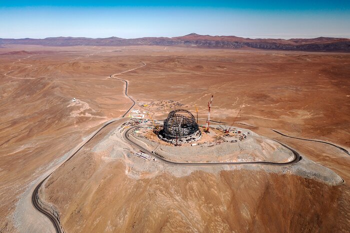 The ELT construction site sits stoically on top of the Cerro Armazones mountain, looking over the vastness of the Atacama Desert from a distance. The familiar round shape typical of telescope domes is clearly apparent in the steel structure of the ELT dome. Roads wiggle around the mountaintop and off into the distance. At the horizon, the flat desert gives way to more mountains, stretching from left to right. The sky above is bright and blue.