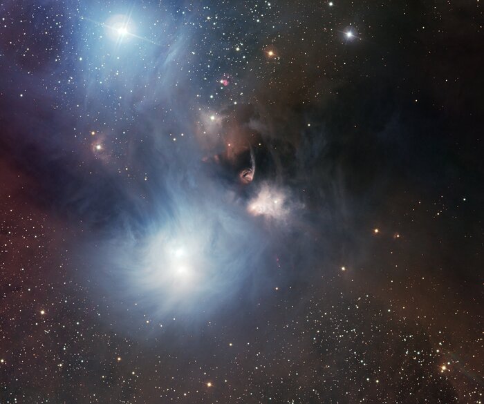 This image shows stars and clouds of gas and dust distributed over a dark background. A prominent cloud of gas and dust can be seen in the central part of the image. It features an amorphous cloud in blue, white, brown colours. Several bright white and blue stars can be seen within the cloud. A handful of stars in the lower part of the picture shine more brightly in yellow and white colours in comparison to the other stars that are distributed uniformly in the image.