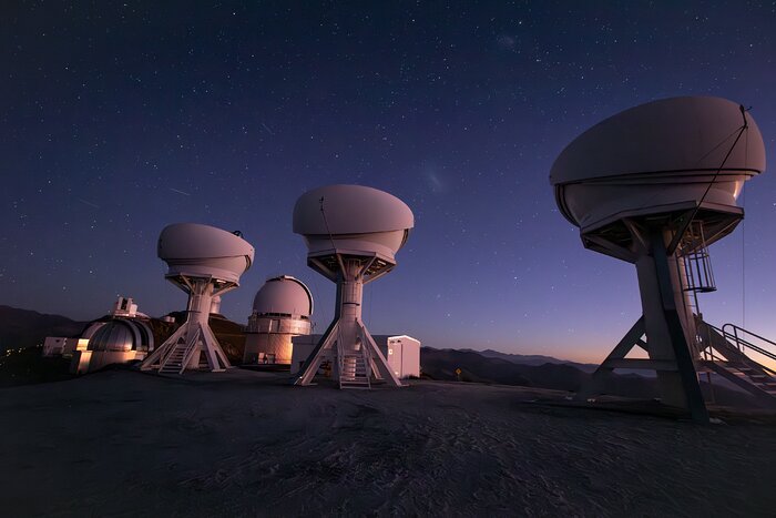 Dieses Foto zeigt im Vordergrund die drei offenen Kuppeln der Teleskope der BlackGEM-Anlage unter einem atemberaubenden Nachthimmel, während im Hintergrund weitere Teleskope des La-Silla-Observatoriums der ESO in Chile zu sehen sind.