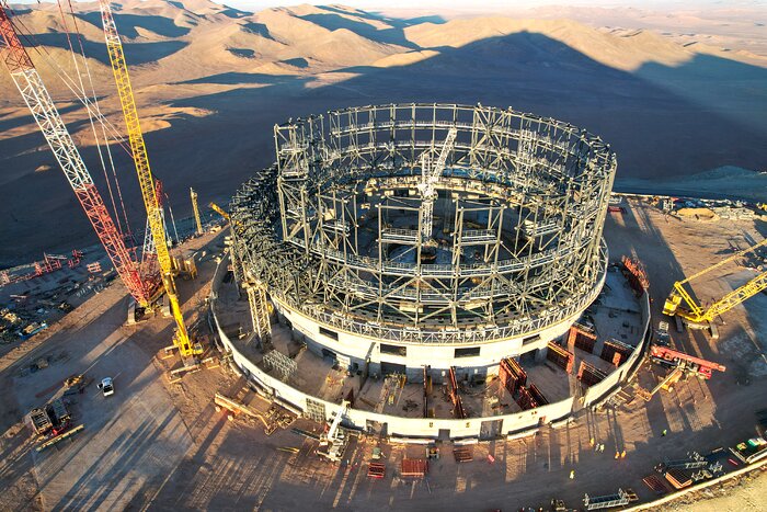 This aerial photograph shows the construction site of the Extremely Large Telescope, ELT, kissed by golden sunlight. The circular structure is made up of steel beams criss-crossing each other, all of which are sat on top of the concrete base. One gets a sense of how big the ELT structure is when comparing it to a white car, parked just left of centre at the left of the image — it is tiny compared to this colossal structure. In the bottom right small ant-like creatures are strolling about — these are the construction workers, dwarfed by the sheer scale of the ELT. Large cranes and other construction equipment are strewn around the outer edges of the structure. Off into the distance behind the construction site the brown, empty landscape with hills and smooth mountains of the Atacama desert stretches out.