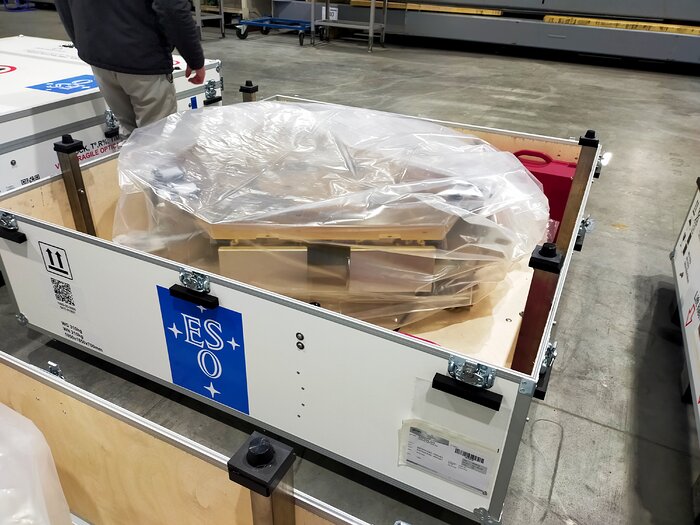 Prominent in the centre of the image, a large white box with the ESO logo — four stars around the letters E, S, O on a blue background — sits on another box. The lid is open, revealing a plastic, almost transparent sheet around a metallic object — a hexagonal mirror segment. A concrete floor and warehouse setting fill the rest of the image.