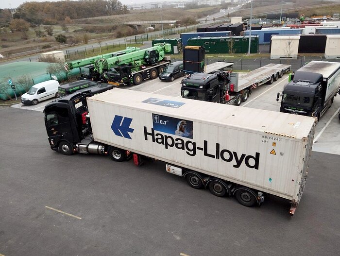 Auf dunkelgrauem Beton ist ein weißer Lastwagen mit der Aufschrift Hapag-Lloyd geparkt, der bereit zu sein scheint, an der linken Seite des Bildes wegzufahren. Direkt dahinter stehen weitere Autos und Anhänger in Parkbuchten. In der Ferne sind verschiedenfarbige Schiffscontainer und braunes Land zu sehen.