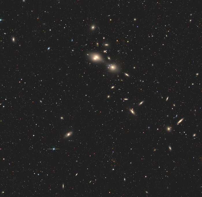 This image shows a large patch of space, dotted with small, distant stars and broken up by fuzzy galaxies spread throughout. These warm, yellow-ish smears of light sit at all angles, some looking more like spirals and some more like clouds. A few galaxies are closer together than others. At the centre of the image is a tiny white dot, which is where the two jousting galaxies can be found.