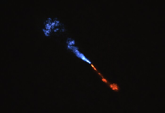 Astronomisches Bild mit schwarzem Hintergrund, das einen stark gebündelten Jet aus einem jungen Stern oder Galaxienkern zeigt. Aus einem zentralen Punkt strömt Materie in zwei entgegengesetzten Richtungen: Nach links verläuft ein bläulicher Jet, der sich in einem leuchtenden Nebel ausdehnt und ausfächert. Nach rechts zieht sich ein orangeroter Jet, der in einer rundlichen, intensiveren Struktur endet. Die Farbgebung symbolisiert vermutlich unterschiedliche Temperaturen oder chemische Zusammensetzungen der ausgestossenen Materie. Das Bild veranschaulicht die gewaltigen Prozesse bei der Stern- oder Galaxienentstehung im tiefen Weltraum.