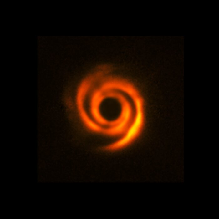 At the centre of this image is a glowing orange swirling pattern, surrounded by complete darkness on all sides. The spiral has two arms that spin around each other, and a large dark hole in the centre. It gives off a fuzzy, uneven light that gently fades into the background.