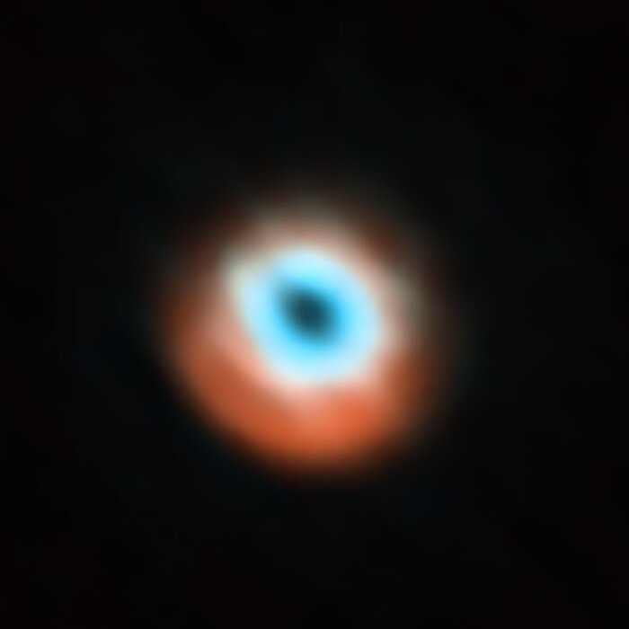 At the centre of this image, surrounded on all sides by complete darkness, is a fuzzy ring-shape. Around the central hole of the ring, it looks bright blue, and towards it's edges it becomes more of a red-orange colour. The red region bulges towards the bottom of the ring, angled slightly to the left and down.