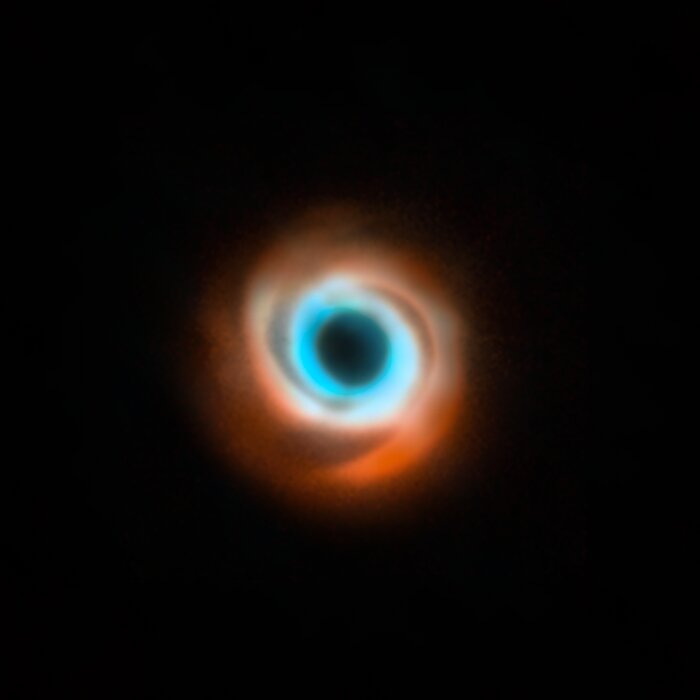 At the centre of this image is a colourful spiral, surrounded by complete darkness. At the outer edges of the spiral, the swirling gas has a appears red, while it's inner regions look like a light blue ring. At the very centre is a dark spot where light has been blocked out by the inertsument that took this picture.