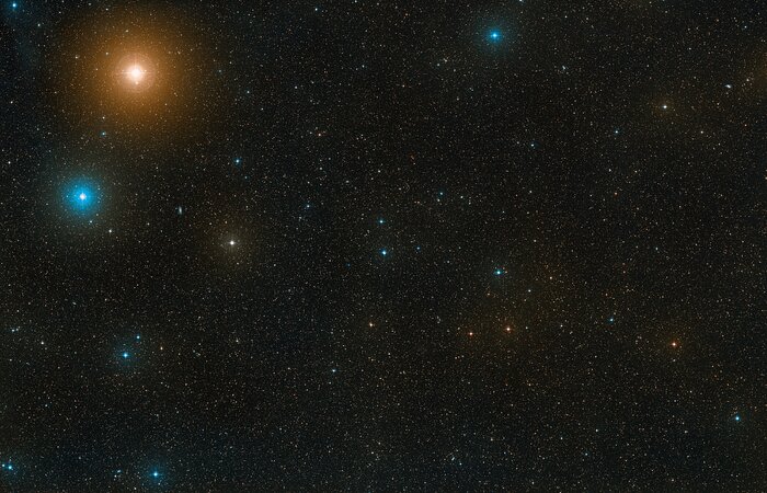 This shows a black, star-filled sky, full of small and dsitant points of white, blue and orange light. Some of the largest stars are on the left hand side of the image, with a bright blue star around two thirds of the way up and far to the left. Another even larger orange point of light is above this star and slightly to the right. At the very centre of the image are two bright, but small-looking, stars which sit very close to each other on the sky.