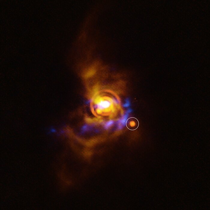 This image shows a colourly patern of oranges and blues against a dark, empty background. At the very centre is a brightly glowing light, surrounded by two orange rings of material and a faint cloud of orange dust, with a bright cone of yellow-ish material pointing to the top left. Underneath this glowing point, curling around it to the right, are faint splotches of blue light. Even fainter orange patches gently fade into the background around the whole system. Just below and to the right of the central light is a circular orange smudge, which has been circled with a white ring to highlight it's presence