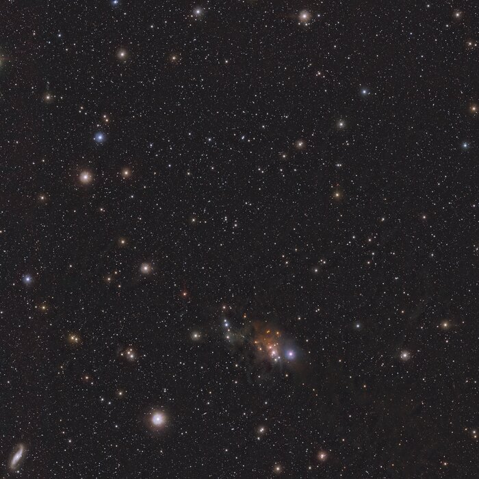 A wide telescopic view of a starry field. There are countless faint stars and a few bright ones in shades of orange, blue and white. At the bottom centre there is a small orange wispy cloud.