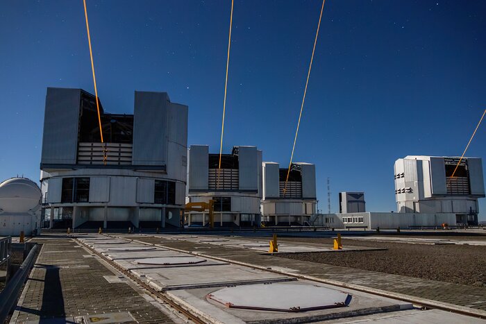 In the spotlight of the photograph are four, big cylindrical structures, with the blue night sky in the background. These grey structures are the domes of the Unit Telescopes, with their shutter doors open. An orange beam emerges from inside of each dome, pointing up into the sky. The observatory site appears bulky, with some pathways and railway tracks. To the left of the image is a smaller, round and white dome.