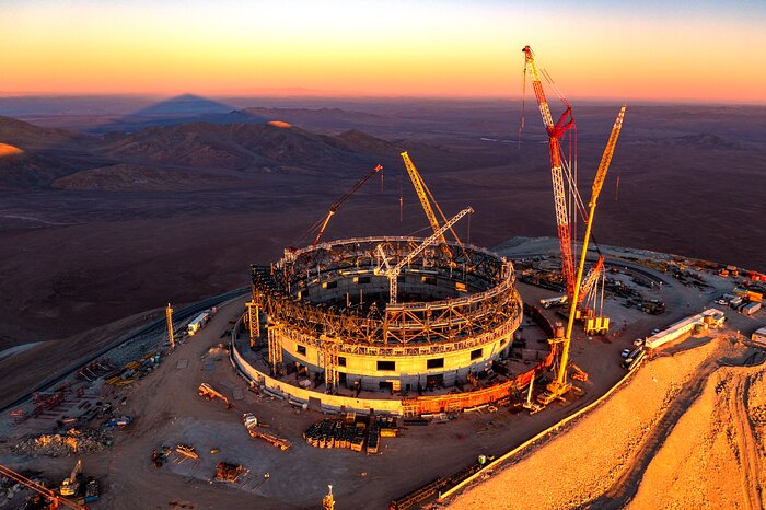 This aerial photograph, taken from a distance in the sky above the construction site, is kissed by the orange light of a setting Sun, streaming in from the right of the image. The ELT is on a flat mountain top and is located in the centre of the image, surrounded by cranes and vehicles. The background is packed full with more brown mountains, some are smooth and rolling, while others are a bit steeper. Above the horizon, the sky moves from deep orange to yellow and white.