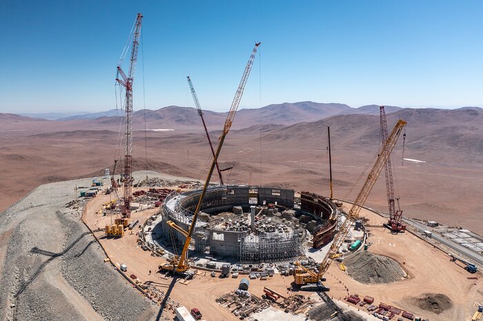 The construction of the circular ELT dome is taking place at a mountain top in the dry, sandy and light brown Atacama Desert. This image shows the construction site, with six cranes surrounding the circular structure of steel and concrete. In the middle of the circle, there is a concrete pillar, which will support the telescope. The sky is clear blue and in the background there are brown mountains.