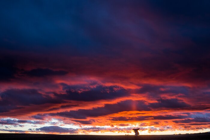 This image is dominated by a large sky filled with blue, purple, and red clouds. Beneath them is one of ALMA’s antennae, which is pointing directly upwards and appears to be illuminated by a beam of red light.