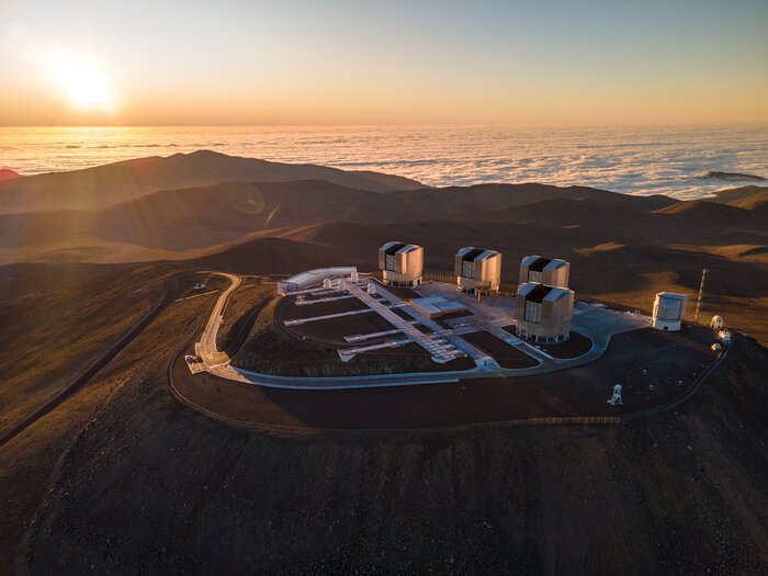 This is an aerial image showing the flattened top of a mountain, dominated by four large metallic cylindrical telescope domes, and a somewhat smaller one to their right. To their left there are four much smaller white domes that look like chess pawns. The desertic landscape is brown and barren, with soft hills, and in the background the Sun is about to set.