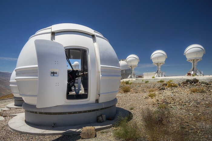 On the left, a white spherical dome occupies the picture; its open doors reveal a small ExTrA telescope inside. On the right, the three white domes of BlackGEM stand in a row on top of pillars. The ground is dotted with small bushes, and the sky is vibrant blue.