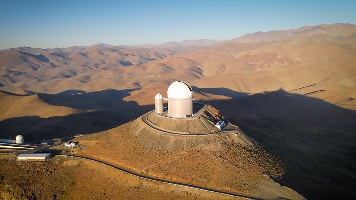 The light blue sky occupies only a small upper part of this aerial picture. The Sun does not appear here, but its white light streams in from the left of the image. Most of the image is filled with brown hills, whose silhouettes are outlined by the shadows they cast upon the ground. At the centre, on the flat peak of a mountain, is the white telescope. The whole infrastructure is made of two similarly shaped buildings, one bigger than the other — cylindrical buildings with a dome at the top. Some roads hug the mountain, and on the left part of the picture are other small white buildings.