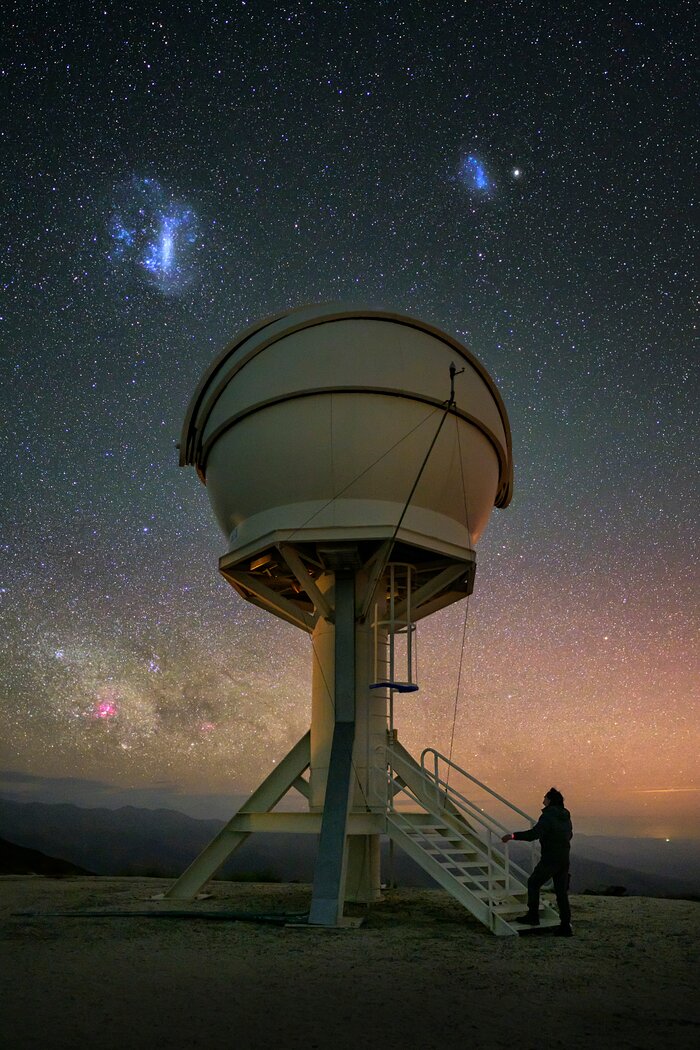 A cream-coloured, spherical telescope dome is suspended above the ground in the centre of this image. On the brownish ground below, a person is stepping onto cream-coloured stairs leading up to the dome. Behind the dome is a colourful night sky, starting orange near the horizon and becoming a dark blue at the top of the image, with white dots sprinkled across. Two bright blue-white cloud looking objects are visible above the dome in contrast to the dark sky.