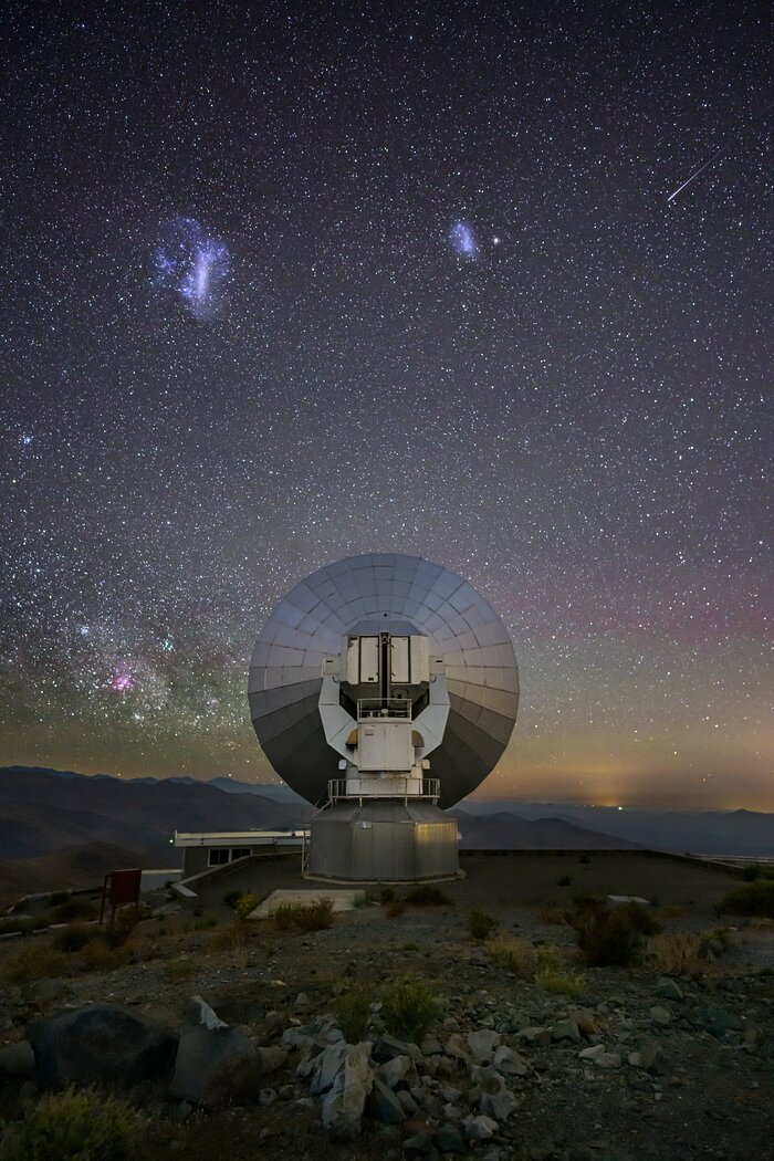 The top half of the image is taken up by the night sky, dotted with thousands of tiny white stars. The sky goes from darker blue at the top to green and orange at the horizon, with faint pink patches in between. In the top third of the image are two white-bluish cloud-like structures and a thin white streak. The steely circular dish of a telescope in the centre of the image reflects some of the colours of the sky. In the foreground we see the rocks and grass on the ground in front of the telescope; behind it, a mountain range in shades of violet and blue stretches into the distance.