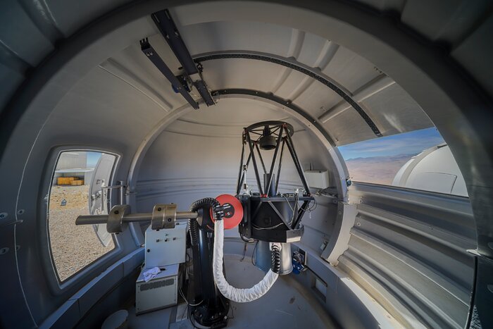 This image is taken within a white dome. In the center is a telescope including a central podium, the main telescope on the right hand side - it is made up of black, diamond shaped rods, and is connected to the central podium both directly and by a white tube - and on the left-hand a bronze counterweight. Behind the telescope, there are white boxes stacked on the floor, a glass door on the left of the dome and a window on the right, through both of which the desert landscape of Chile and other domes can be seen.