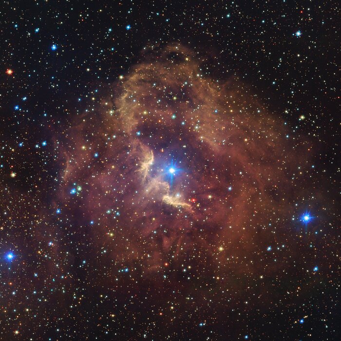 This image shows the GUM 41 nebula, which takes up most of the frame. It is a reddish-brown, circular-shaped nebula with a bright central star. There is a second bright star outside of the nebula towards the bottom right of the image, circled by some additional, fainter reddish-brown clouds. The background of the image is dotted with stars of many colours and sizes.