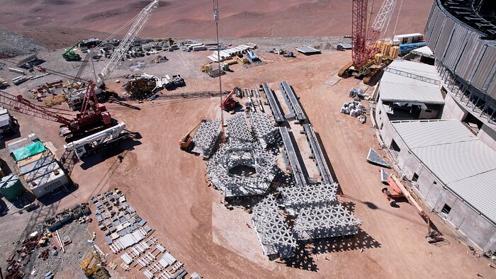 Against the landscape of the Chilean desert, the foreground of this image shows multiple pieces of large, white, honeycomb lattice structures, with the piece central in the image being hexagonal in shape. The lattice pieces are surrounded by construction instruments, cranes, and trucks, and on the right of the image, the large dome of the ELT is partly visible.