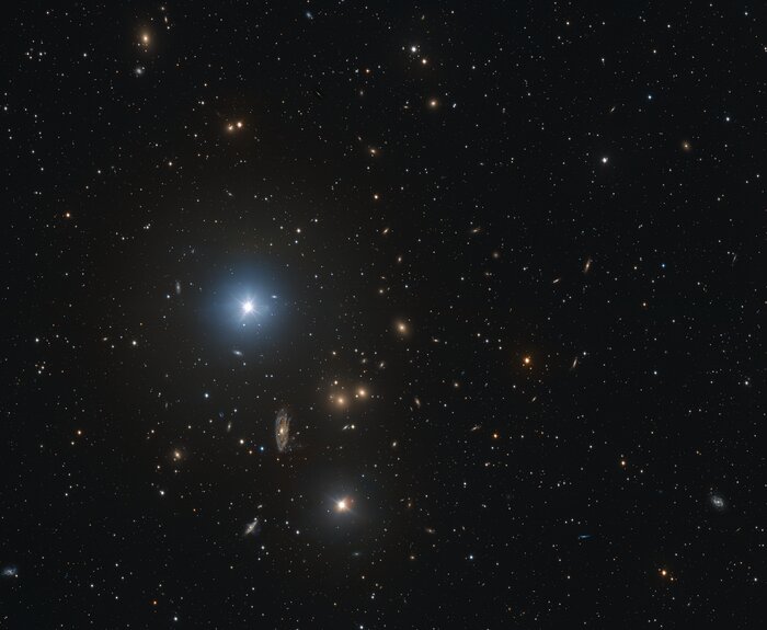 The image shows a colourful backdrop of stars and galaxies against the black cosmos. There is a particularly large and bright star slightly left of the centre, and below this, a clear spiral galaxy. The spiral galaxy appears slightly smudged.