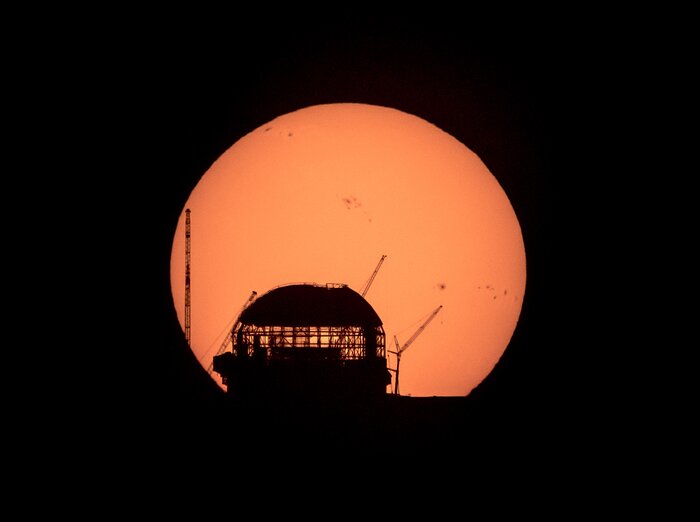 A huge orange Sun, showing some small dark spots towards the top and on the right side, contrasts against a black background. In front of the orange Sun, but only a quarter of its size, the shadow outlines of a dome show in black. It is a work in progress, as the top is dark, the middle shows light piercing through its structural beams, and the bottom is dark too. Three cranes and a high pole are standing near the dome.