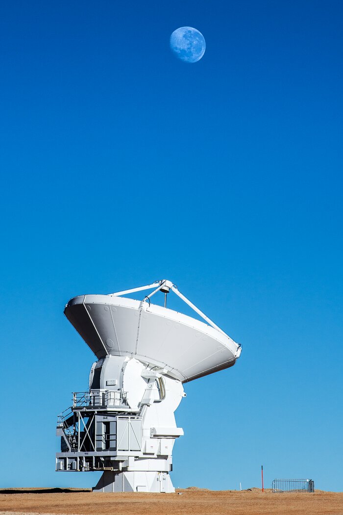 A white large antenna stands alone in the Atacama desert. The background is of a striking blue colour that ranges from darker at the top to light blue at the bottom. A full Moon is faintly visible on the upper part of the image, to the right, and the antenna faces its direction almost perfectly.