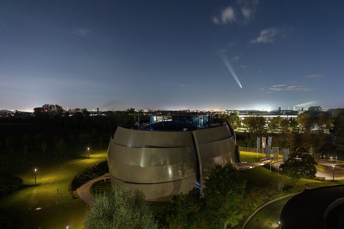 An oval, futuristic-looking building stands in a dim-lit garden area: the ESO Supernova Planetarium and Visitor Centre. The picture was taken late in the evening, when the night had just started and the sky still had a dark-blue hue. The sky is mostly clear, except by a couple of small clouds and a comet with a long tail positioned very close to the horizon. The horizon is dimly lit by city lights.