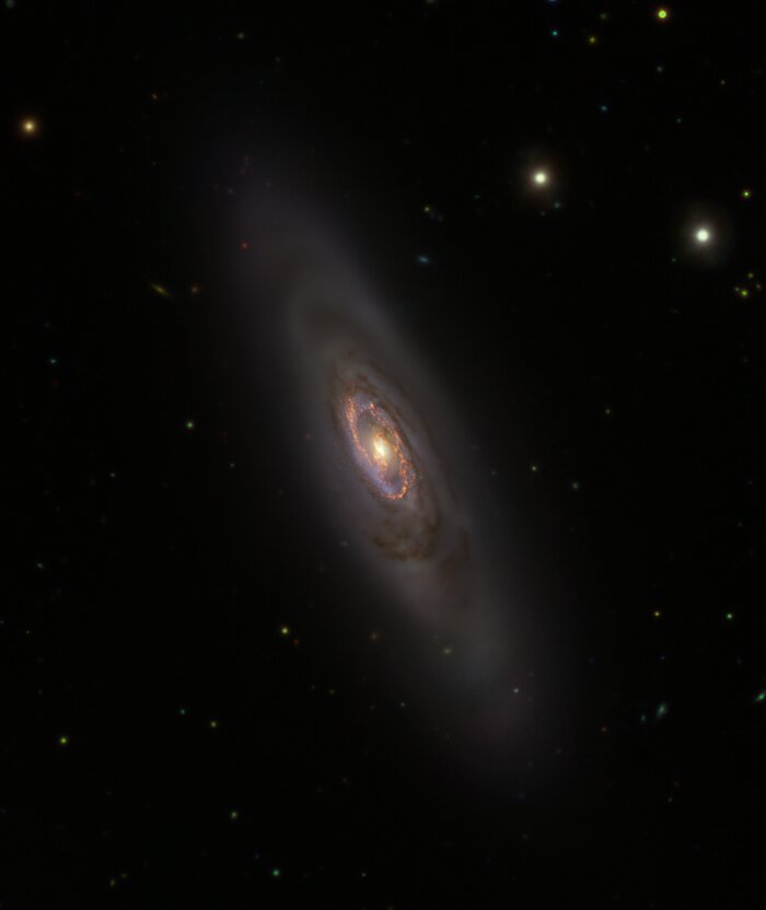 The picture shows a dark region of space, dominated by a large galaxy and other single stars visible in its surroundings. The galaxy is elongated like an ellipse and appears in a faint white colour. The centre of the galaxy is highlighted in blue and golden colours, which represent young stars and clouds of gas respectively.