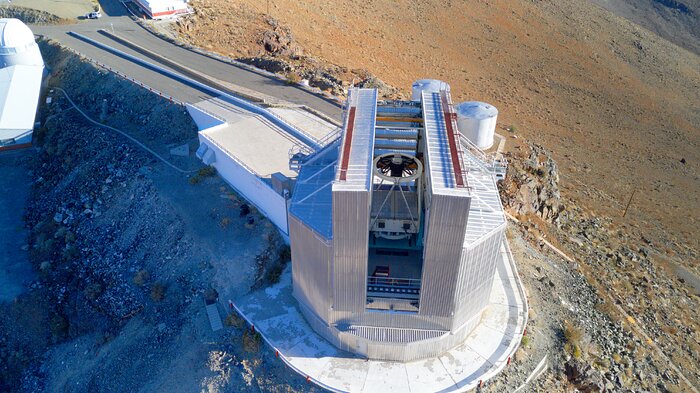 The image shows the edge of a cliff in a barren, arid landscape, seen from a bird's-eye perspective. Along the edge of the cliff there is a wide, short road that finishes in front of a silver building. This building is several stories tall and has a distinct shape, overall being octagonal with two main towers that elevate in the shape of an “H”. In between these towers there is a white cylindrical structure; the supporting structure holding the main mirror of the New Technology Telescope.