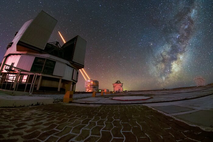 Against the backdrop of a dark night sky full of stars and the Milky Way, a large telescope stands to the left. Two telescopes are in the background, the left one shooting four bright orange laser lights towards the sky. The scene has a brown-pinkish hue. The night sky is showing behind the telescopes, with a yellowish-glow at the horizon, strongest in the centre of the image and more bright blue to the right, where the Milky Way is.