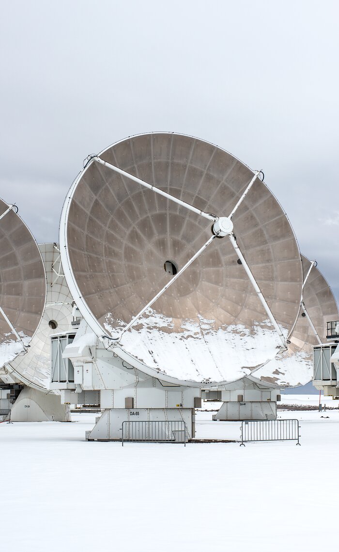 A radio telescope stands in the middle of the image, taking up most of the space. It stands in white snow, against a completely clouded, light gray sky. The huge dish of the antenna is aimed to the right, its metallic brown-gray inside curve showing, the bottom third of it covered in white snow. Behind the antenna, parts of other similar antennas are showing.