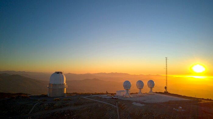 Four telescopes stand on top of a hill. The telescope on the left is cylindrical and larger than the other ones. The three remaining telescopes stand very close to each other. They are smaller and their shape resembles a short lollipop, or a mushroom. All telescopes are white. The sky is of an intense blue, and the whole picture is bathed in the orange colours of the sunset. In the background, the Sun is about to set, and other mountain ranges appear faintly in the distance.