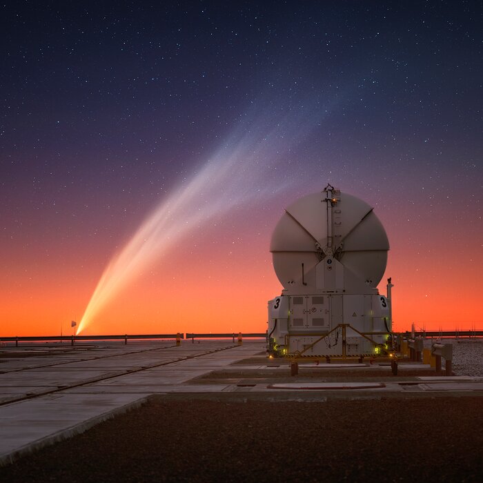 A sunset photograph at an observatory. The colour of the sky changes from blue, almost purple, to golden at the horizon. There’s a white closed telescope dome with a spherical top. To its left, the comet stretches diagonally across almost the entire image, as if it had been painted on the sky with a gigantic brush. The tail of the comet is white and full of intricate filaments.