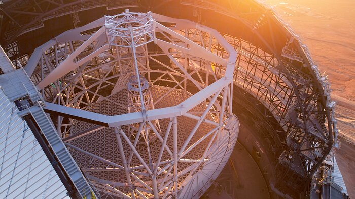 The drone image shows the inside of the Extremely Large Telescope as seen from above, sideways through a big aperture in the roof of the telescope dome. In the middle of the dome stands the telescope's altitude structure. Constructed from sturdy beams, it is white and has the shape of a long decagonal prism. Orange light is shining through the lower part of the steel frame of the dome surrounding it, which is uncovered.To the left of the image, next to the aperture, are stairs to climb the dome roof.